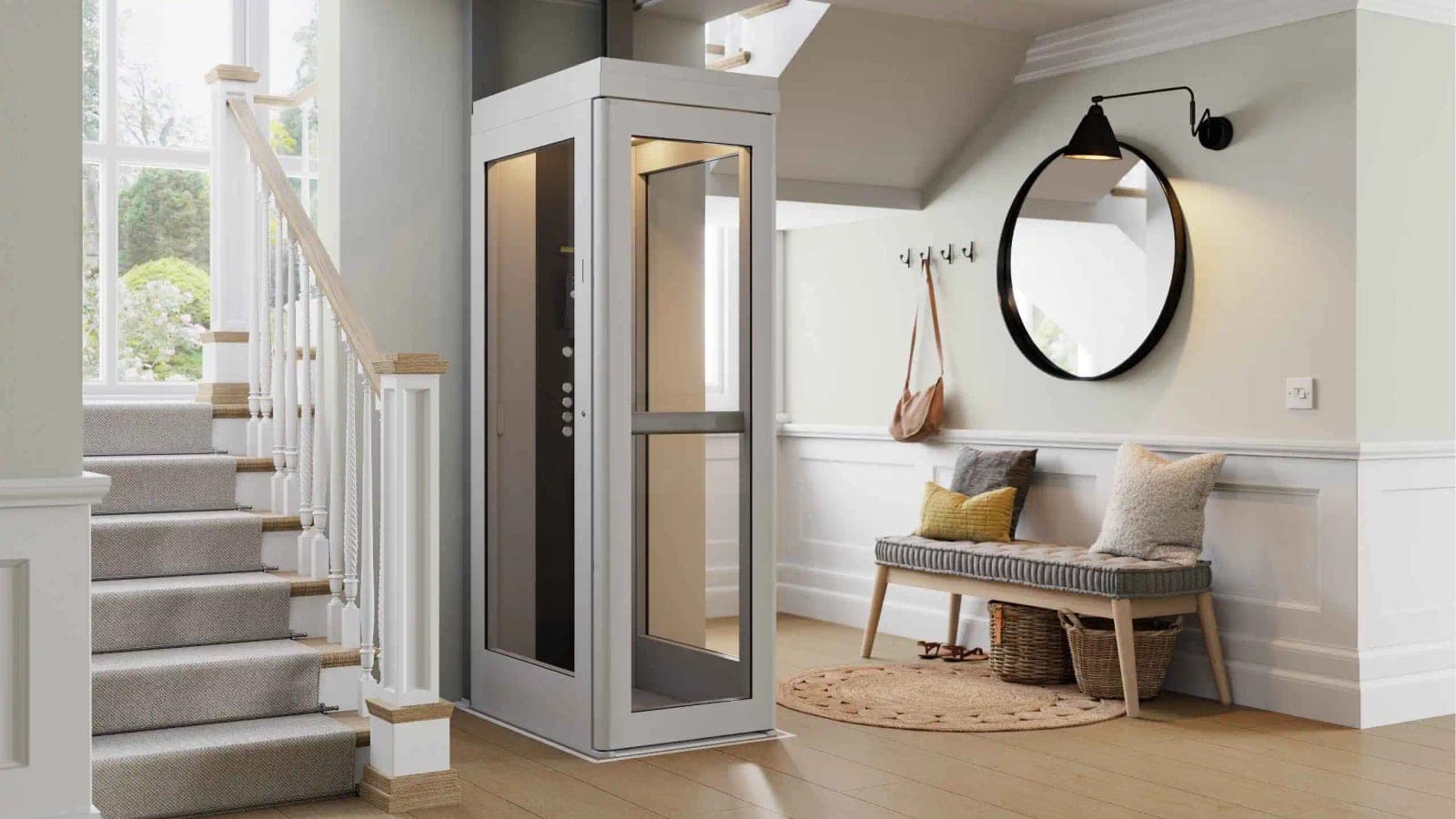 Bruno Connect Compact home elevator in a stair hall beside the staircase: a free-standing single-user cab with a light gray frame, clear front and side panels, a horizontal handle bar across the front and control buttons on the inside wall.
