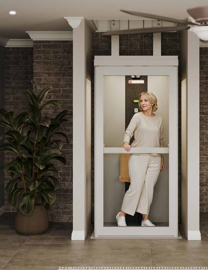Spire Home Elevator with a light-colored frame and a full-height glazed door with a horizontal handrail, a woman standing inside the cabin beside a folded tip-up seat, in a room with a brick wall and a tiled floor