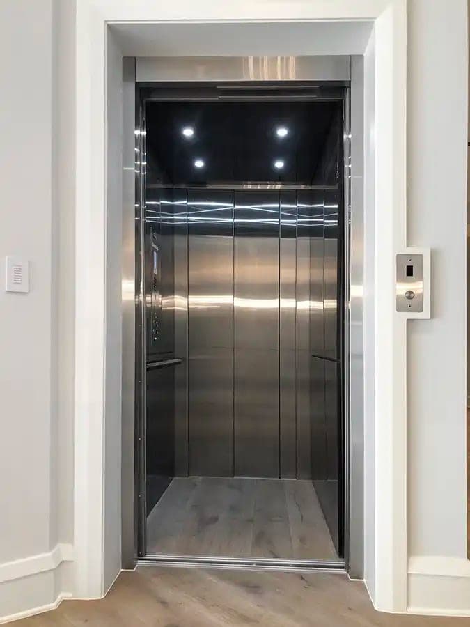 Savaria Eclipse home elevator seen from the landing, with its stainless steel automatic sliding door open onto a stainless steel cab with four recessed ceiling lights, side handrails, a wood floor and a closed rear door for straight-through access. A hall call button is on the wall to the right of the entrance.