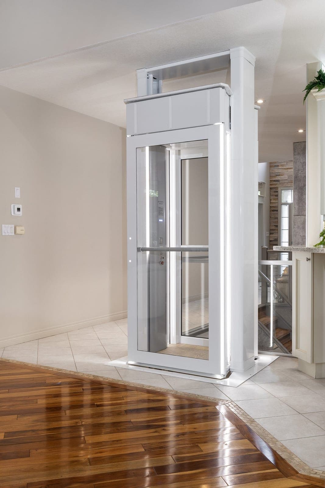 Savaria Luma through-floor lift in a home, seen from the front corner: a white enclosed cab with clear panels and a full-height clear door with a horizontal handrail across it, the drive box on the cab roof, and the two white rails rising to the ceiling, standing on a tiled floor beside a wood floor.
