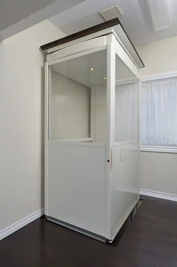 Savaria Telecab through-floor lift at the upper level of a home, seen from the front corner: a white enclosed cab with solid lower panels, clear upper panels, a closed door with a small handle, and the floor plug on the cab roof finished in dark wood, standing on a dark wood floor beside a window.