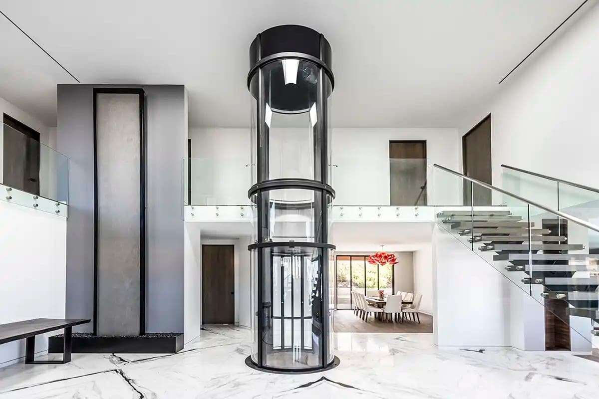 Savaria Vuelift round home elevator with a black frame and clear curved panels, car at the lower landing, standing in a two-story hall beside a mezzanine and an open staircase.