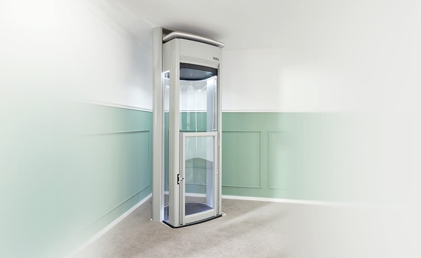 Stiltz Duo Alta through-floor homelift with a light grey frame, curved clear side panels and a half-height door, standing against the wall of a carpeted room