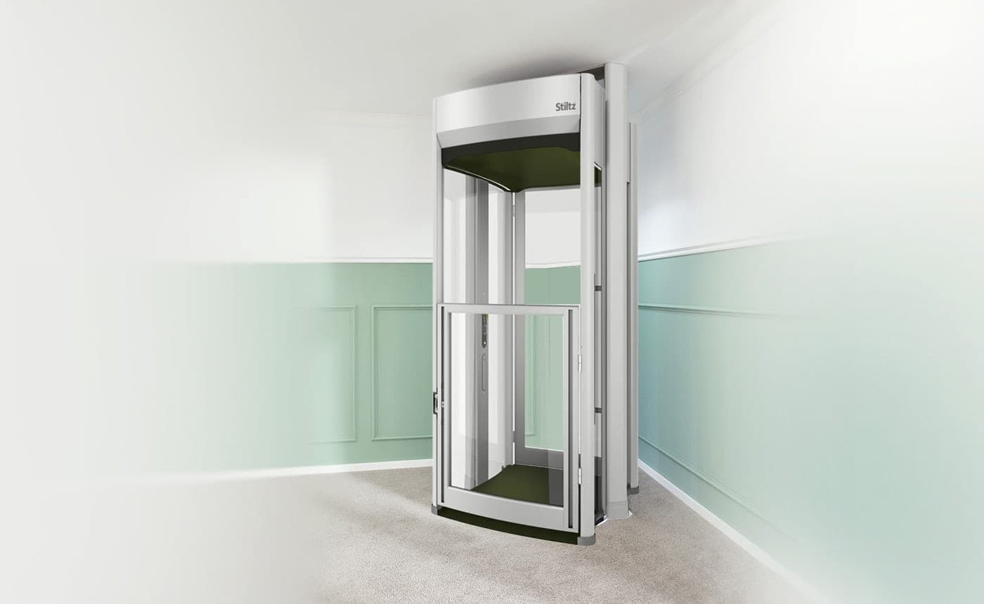 Stiltz Trio Alta homelift with glass car walls and a hinged half-height door, standing on a carpeted floor in the corner of a room, with its vertical guide rails behind the car.