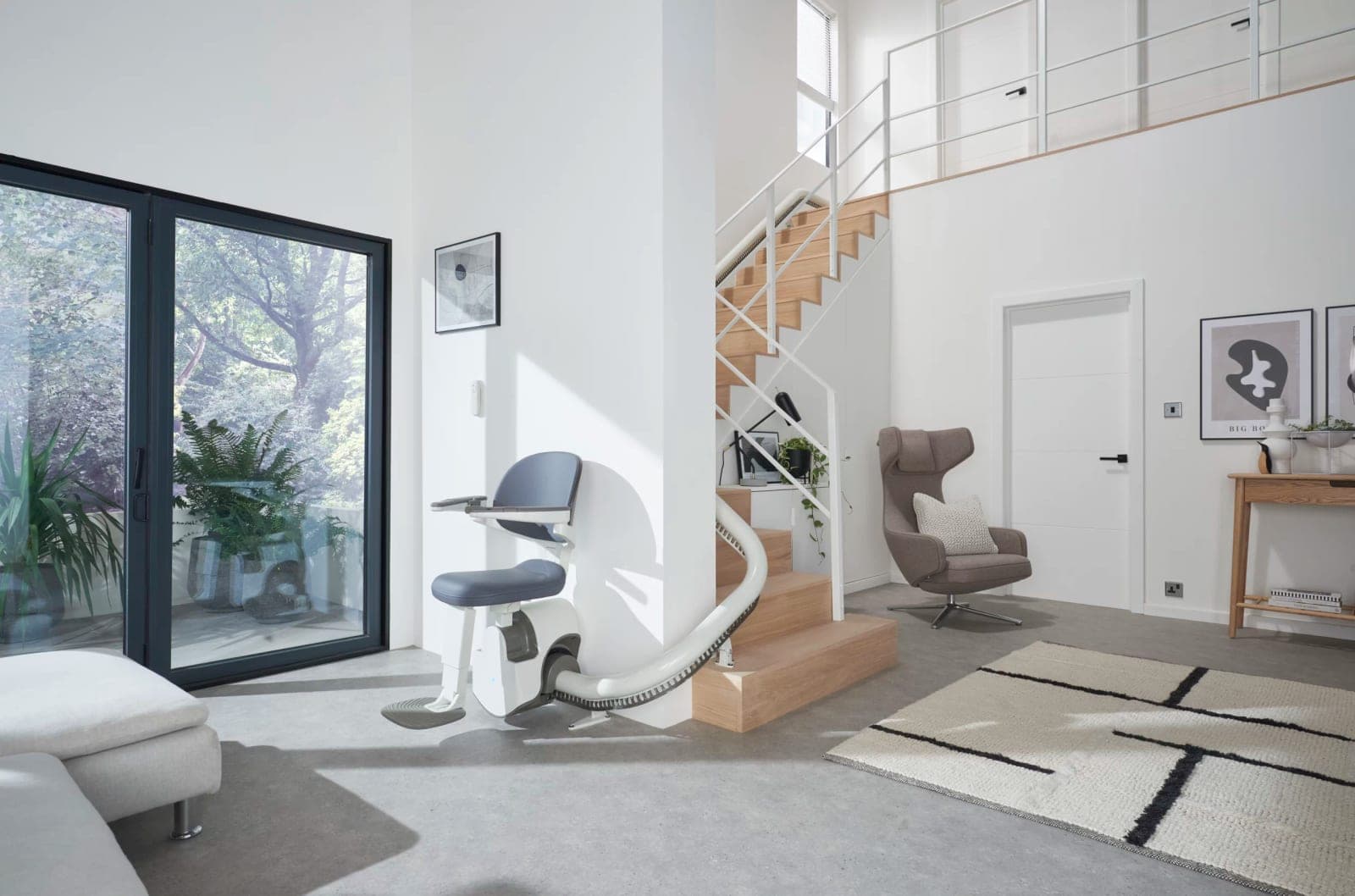 Access BDD Flow X stairlift parked at the foot of a wood staircase in an open living room: an empty chair with a white frame, blue-gray seat and backrest, armrests and footrest lowered, on a white rail that bends out from the bottom step and runs up the stairs.
