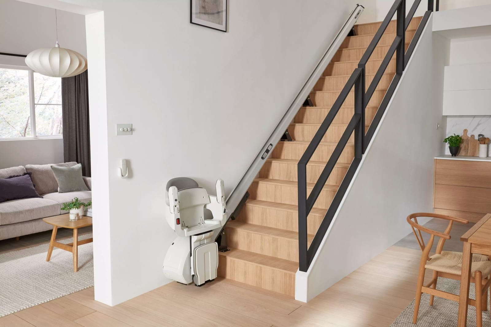 Access BDD HomeGlide straight indoor stairlift parked at the bottom of a straight wooden staircase in a home, with its white seat, armrests and footrest folded up and its aluminum rail fixed to the stair treads and running up the stairs.