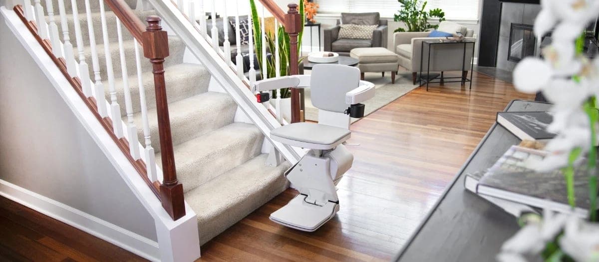 Bruno Elan SRE-5000 straight indoor stairlift parked at the bottom of a carpeted straight staircase in a home, with its light gray seat, armrests and footrest unfolded and its rail running up the stair treads.