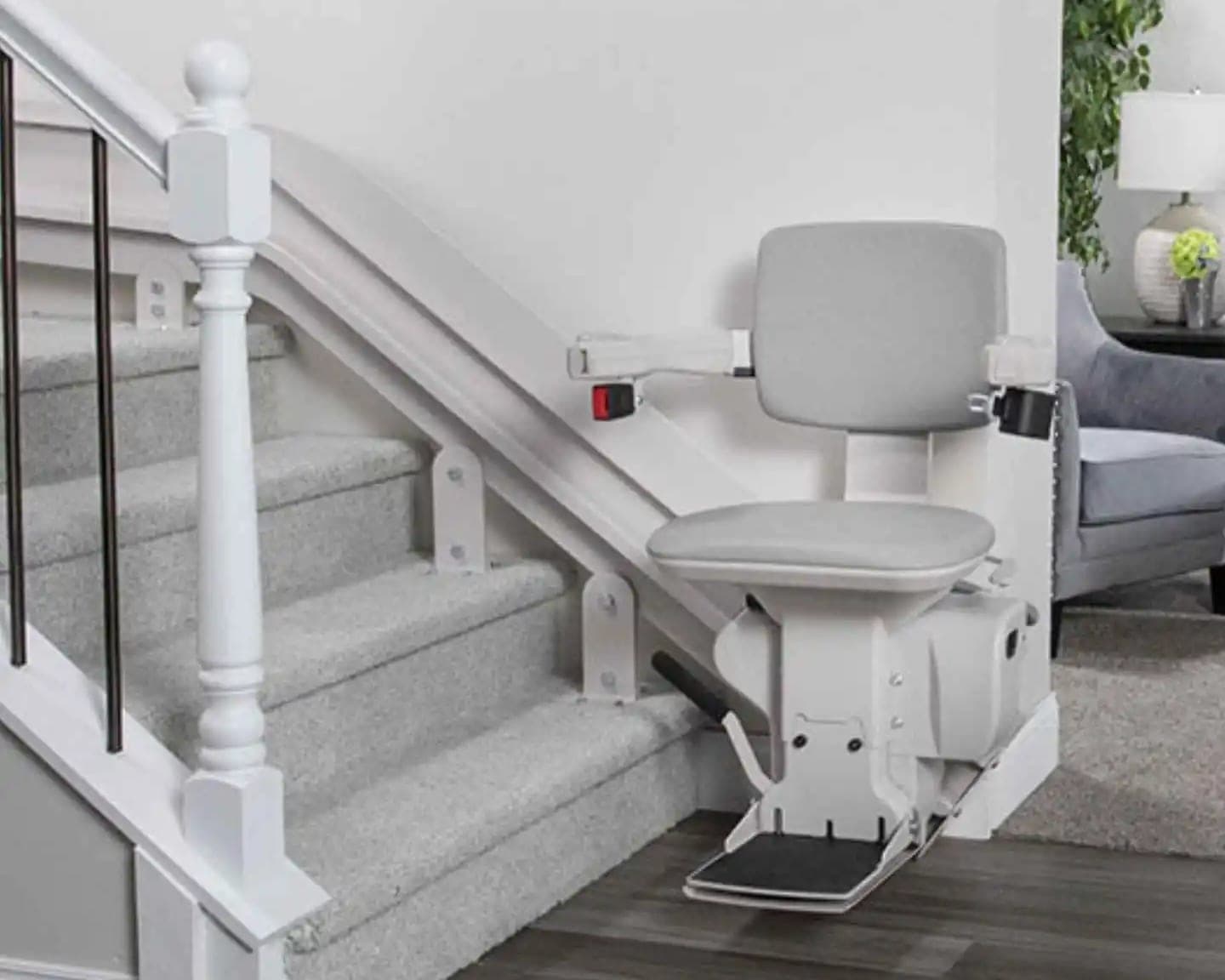 Bruno Elite Curved CRE-2110 indoor stairlift parked at the bottom of a carpeted staircase in a home, with its light gray seat, armrests and footrest unfolded. Its white rail is fastened to the stair treads on brackets and bends toward the landing near the top of the flight.