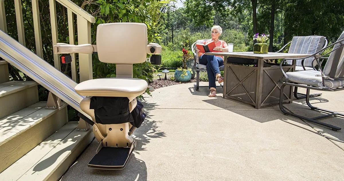 Bruno Elite straight outdoor stairlift, model SRE-2010E, parked at the bottom of a painted wooden deck stair on a concrete patio, with its tan seat, armrests and footrest unfolded, its black light-weight cover stowed below the seat and its aluminum rail running up the steps; a woman reads at a patio table in the background.