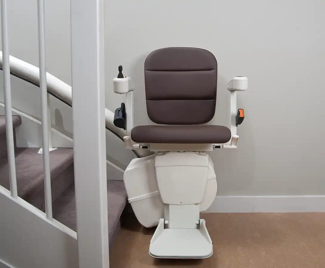 Handicare Freecurve curved stairlift parked at the bottom of a carpeted staircase that curves up to the left. The seat and backrest are brown, the carriage and folded-down footrest are cream white, and a joystick is on the left armrest. The single-tube rail runs up the stair behind a white balustrade.