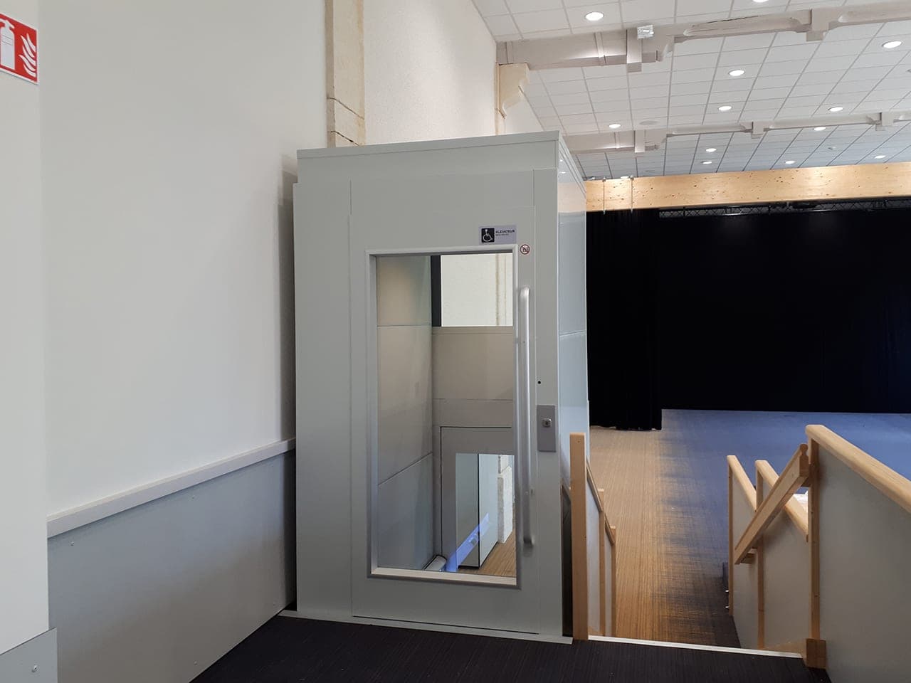 White Cibes Primo platform lift in its own enclosed shaft, with a full-height landing door that has a glass vision panel, beside a staircase in a school building.