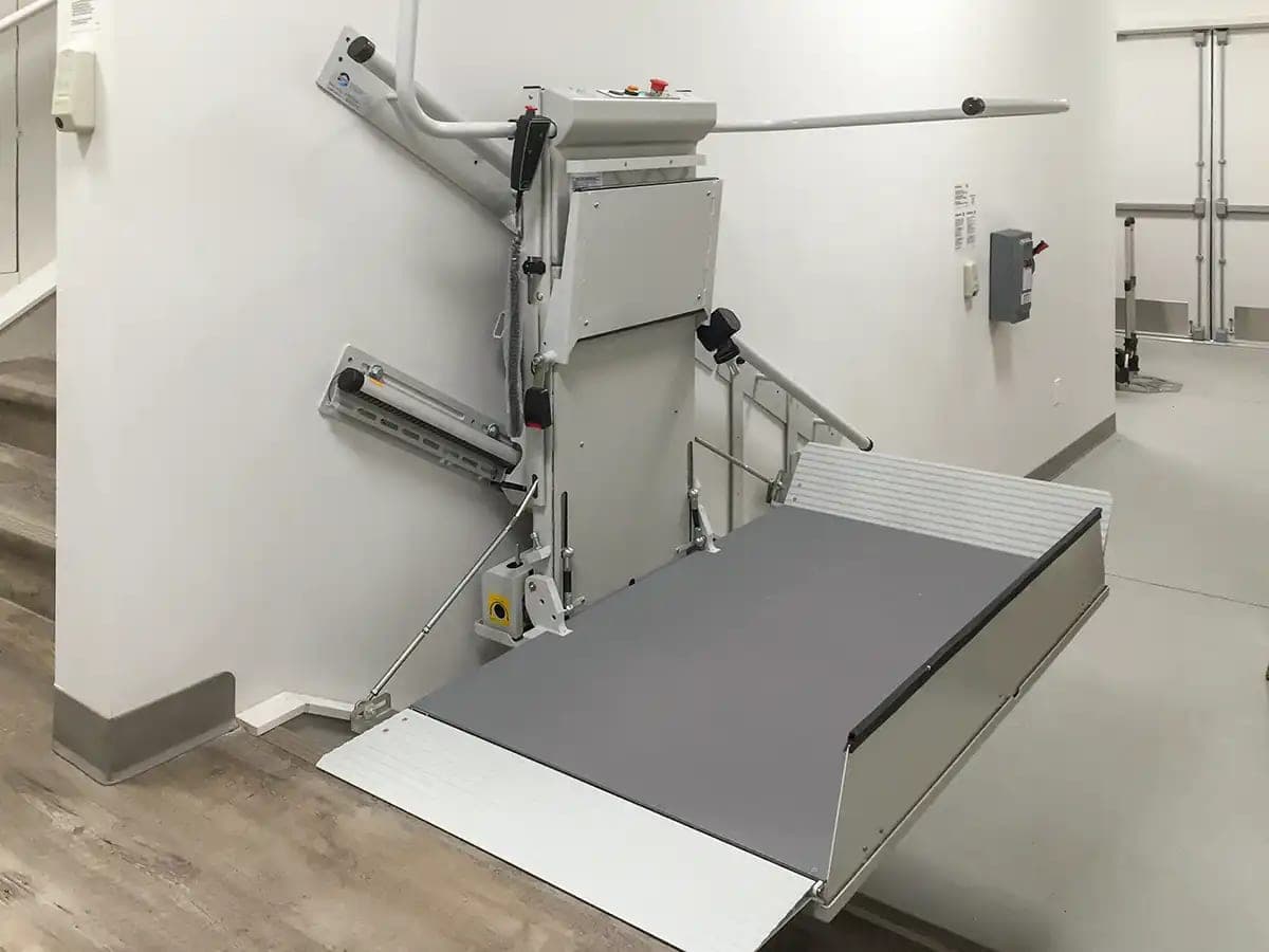 Savaria Delta inclined platform lift in an indoor stairwell: the gray wheelchair platform unfolded with its entry ramp down, the drive unit with its control panel and hand-held pendant, and the upper and lower straight rails fastened to the wall.