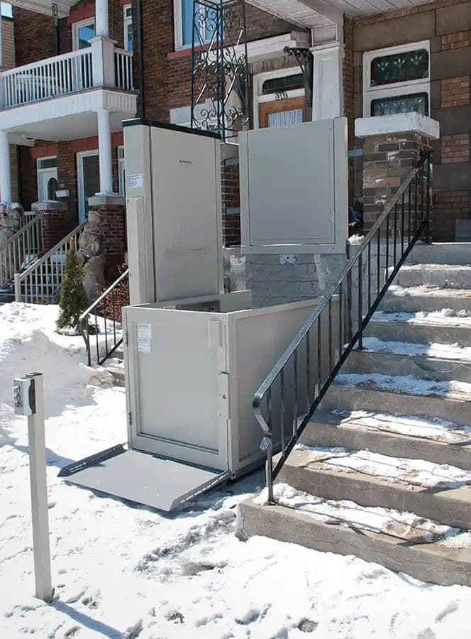 A Savaria Multilift vertical platform lift beside the concrete front steps of a brick row house in winter. The tower, the platform with its side panels and gate, the lowered access ramp and the top landing gate at the porch are all visible.