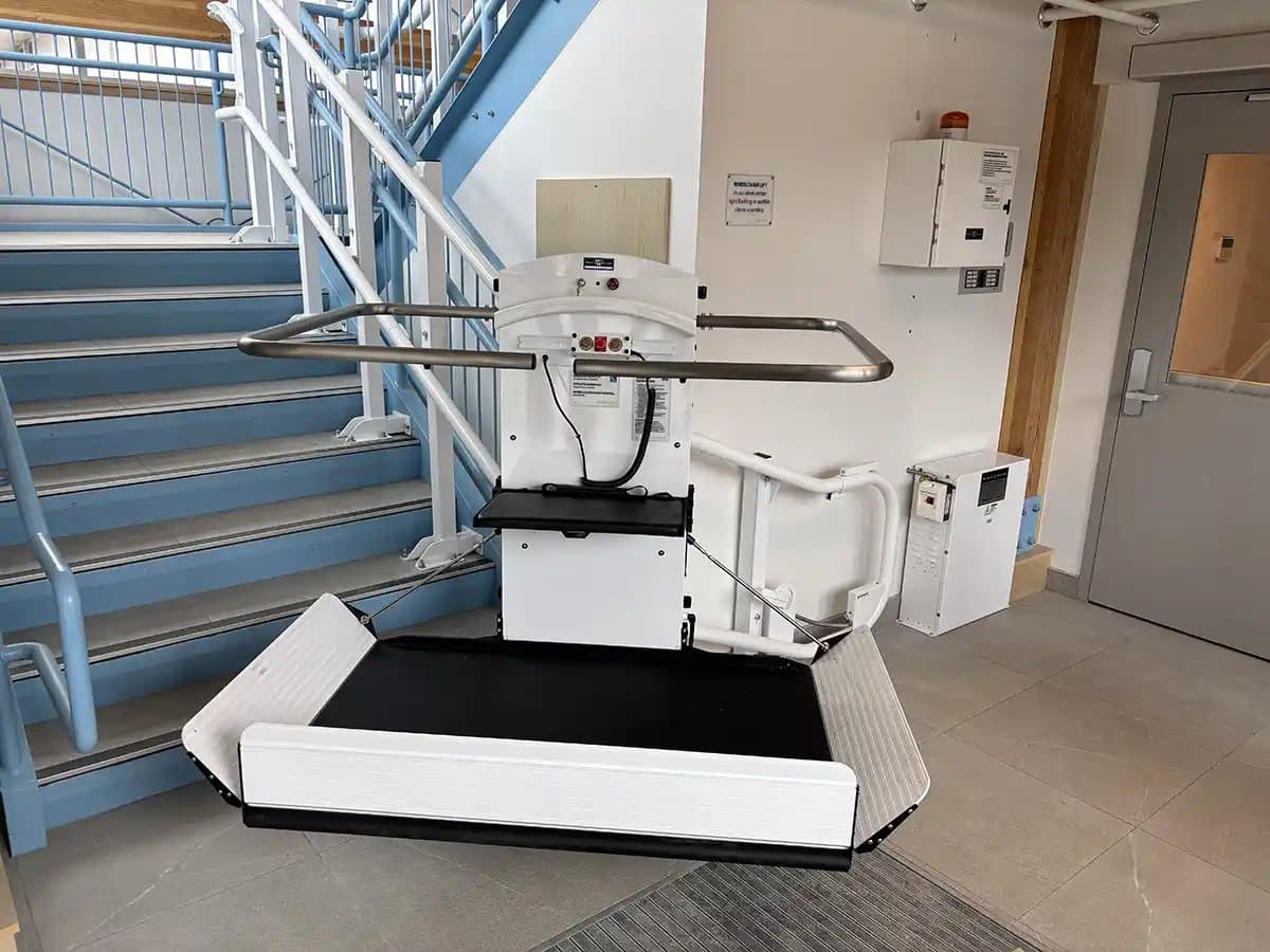 Savaria Pegasus inclined platform lift at the foot of a commercial staircase: the white carriage with its control panel and pendant, the black platform unfolded with both side ramps raised and the safety arms down, and the two white guide tubes bending around the end of the stair.