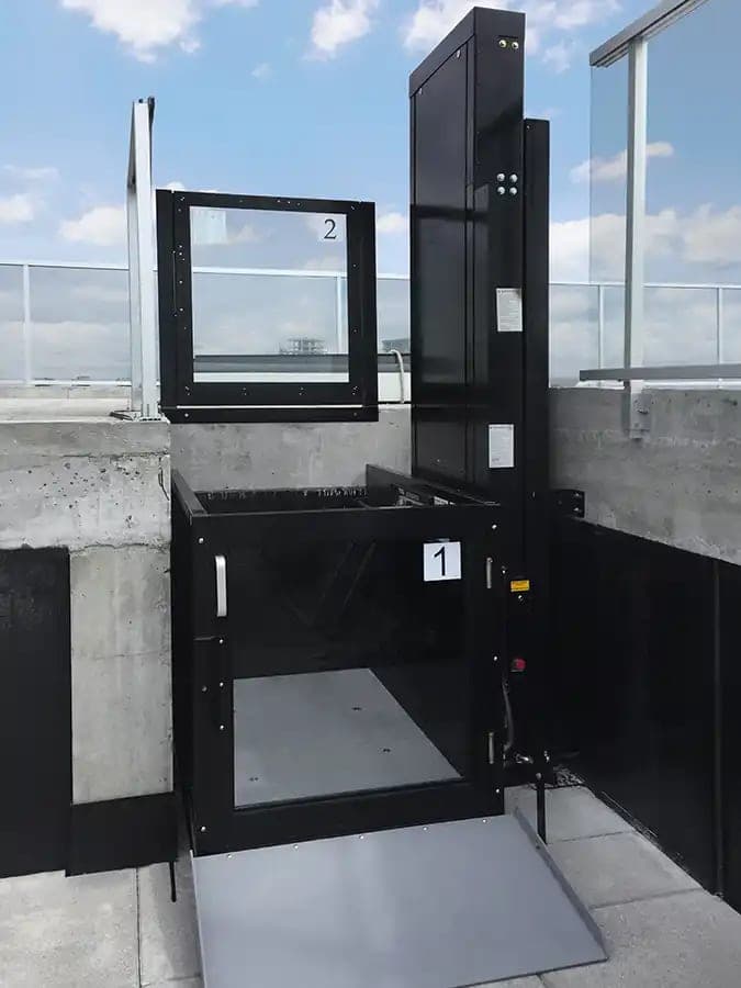 Savaria V-1504 Standard unenclosed vertical platform lift in a black finish on a building rooftop. The platform is at the lower landing with its gate closed, the drive tower is on the right, an access ramp leads onto the platform, and the upper landing gate with a glass insert stands at the raised level behind it.