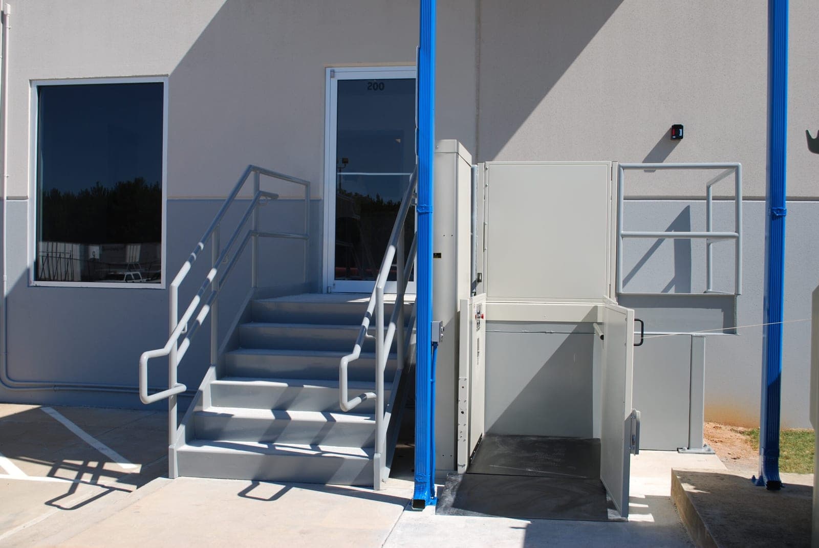 Cibes Symmetry VPL-UL unenclosed vertical platform lift in a light gray finish beside the entry stairs of a commercial building. The platform is at the lower landing with its gate open, the machine tower is on the left and the upper landing gate stands behind the platform.